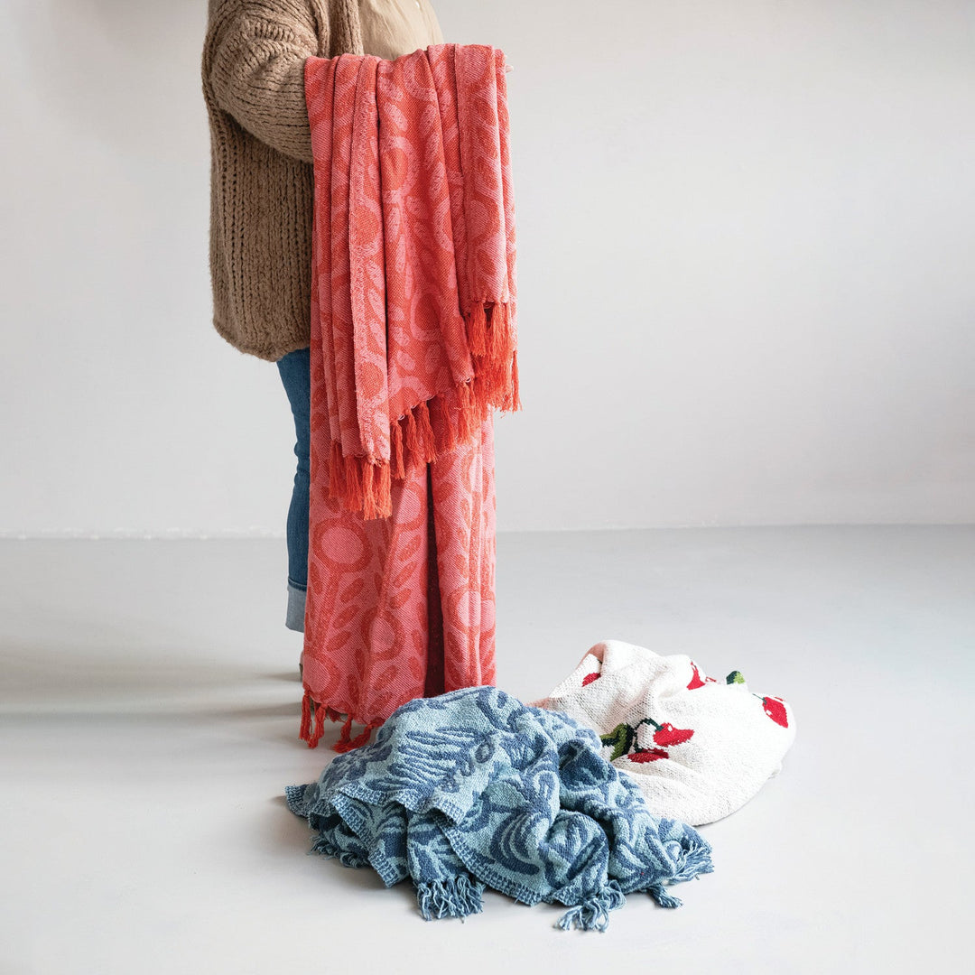 Pink floral jacquard throw styled held up showing drape and fringe with more throws piled below in a Portland studio - Whats New Furniture