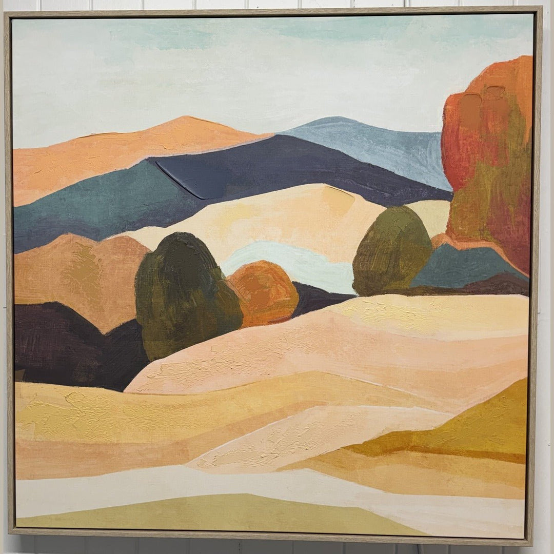 Framed landscape painting of layered hills in a light wood floater frame, hung in the What's New Furniture showroom