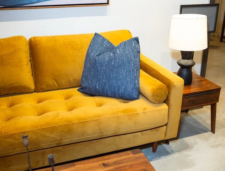 A mustard velvet sofa with a blue pillow beside a wood side table and lamp in the Cedar Mill showroom