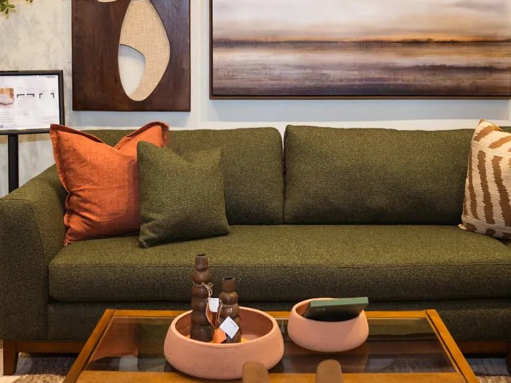 An olive green sofa with rust and green pillows and a glass coffee table under abstract wall art in the Cedar Mill showroom