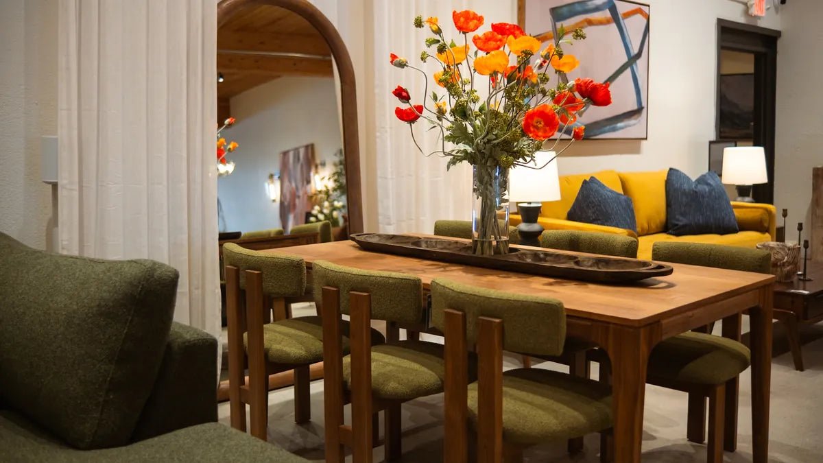 A walnut dining table with orange poppies and green upholstered chairs, with a mustard velvet sofa beyond, in the Cedar Mill showroom