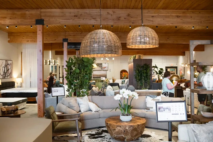 Shoppers on our Cedar Mill showroom floor under rattan pendant lights, with product cards on stands by the sofas - Whats New Furniture Portland OR